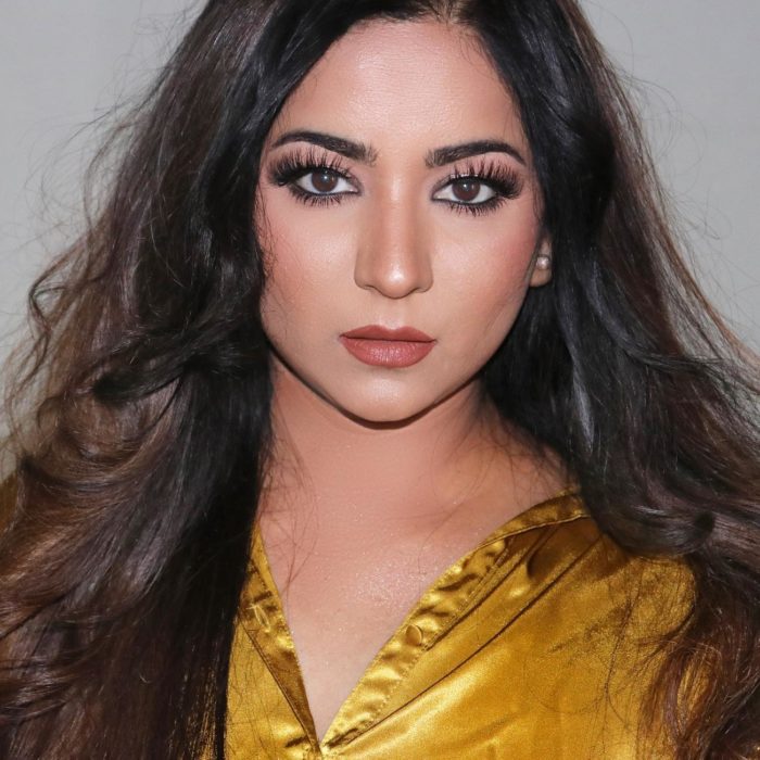 Smitha Deepak looking into the camera wearing a gold top in front of a gray background