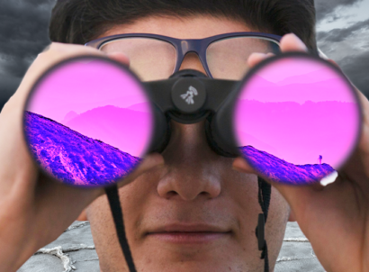 man in a black and white dry landscape looks through binoculars. In the binoculars' lenses, a colorful mountain ahead to represent