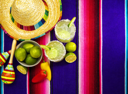 This is a photograph of two modern margarita glass with a rim of salt surrounded by fresh cut limes and chilis on a colorful striped mexican blanket next to a sombrero