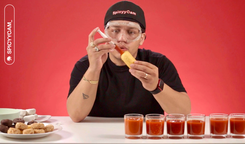 TikToker Spicy Cam sitting at table injecting hot sauce into food item while wearing lab goggles.