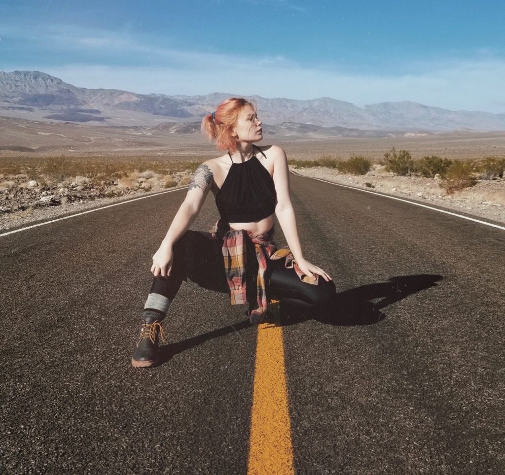 Jo Steel kneels in the middle of an isolated road in a desert/mountain landscape