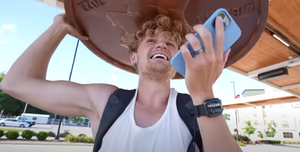 An excited Ryan Trahan holds a giant penny object above his head while talking on his mobile phone.
