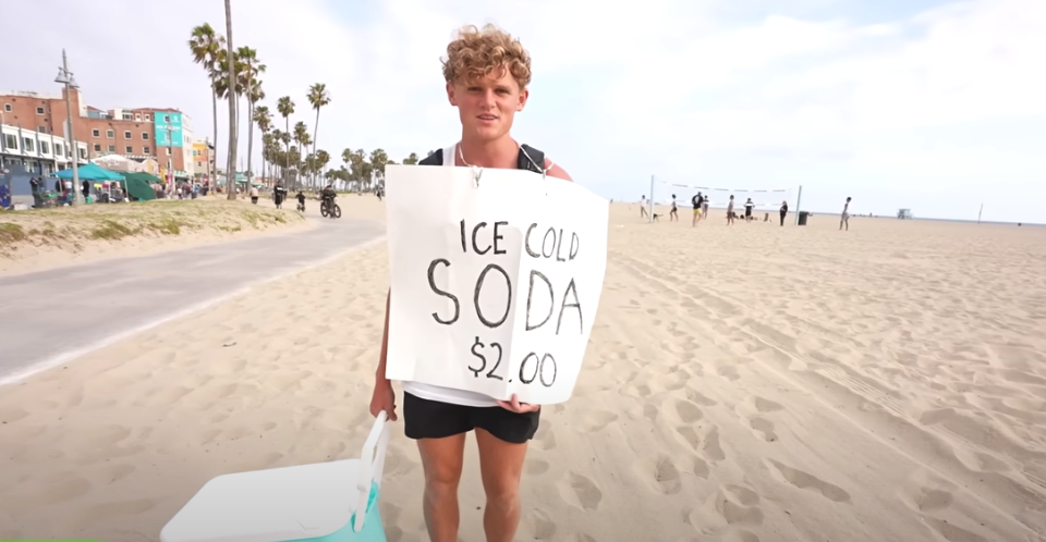 YouTube creator Ryan Trahan stands on the beach holding a sign that says Ice Cold Soda $2