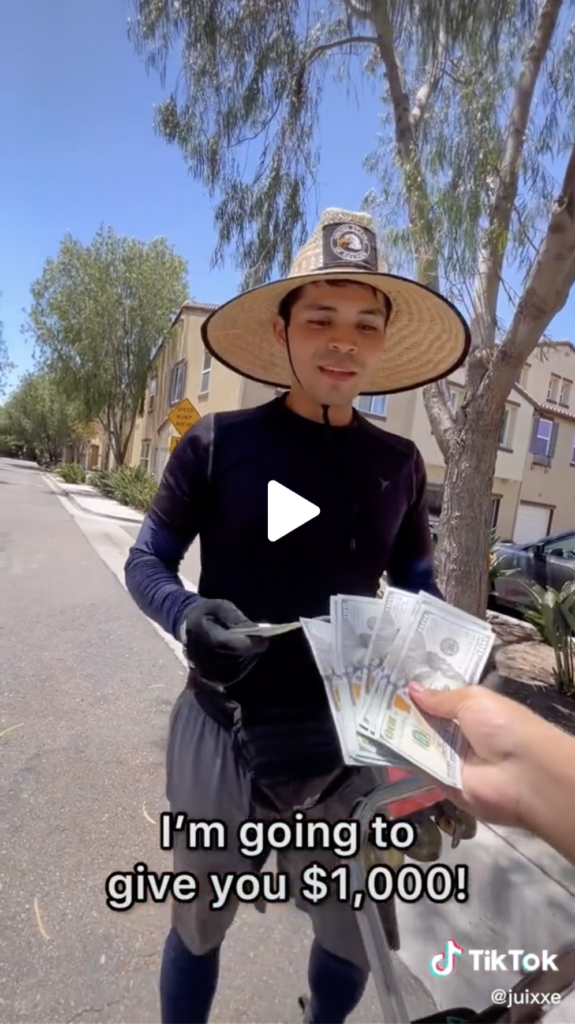 Male street vendor wearing a hat and black long-sleeve shirt outside is given $1,000 from creator Juixxe