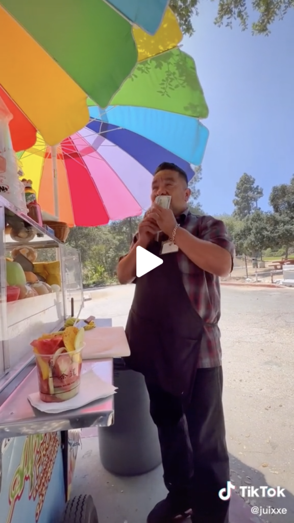Male street vendor stands under rainbow umbrellas outside kissing money gifted to him by TikTok creator Juixxe