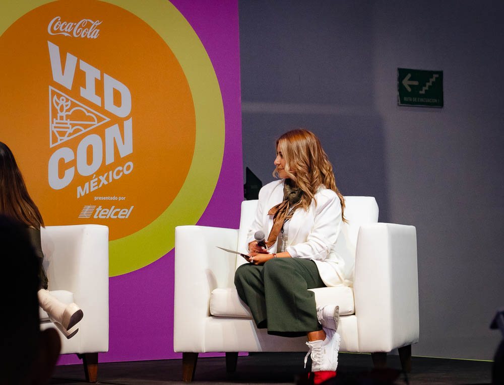 Female sits in white arm chair on stage at VidCon Mexico conference. 