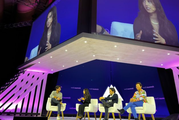 Four panelists sit on stage beneath a large screen that shows a female panelist speaking.
