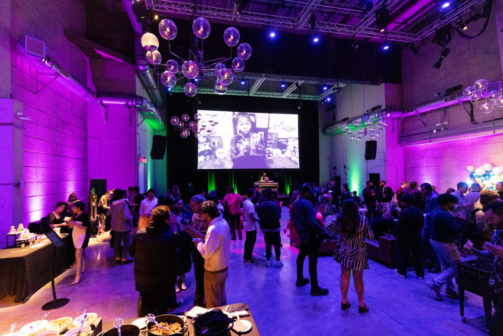 A crowded event space with a big screen playing a black and white video with people standing eating and drinking