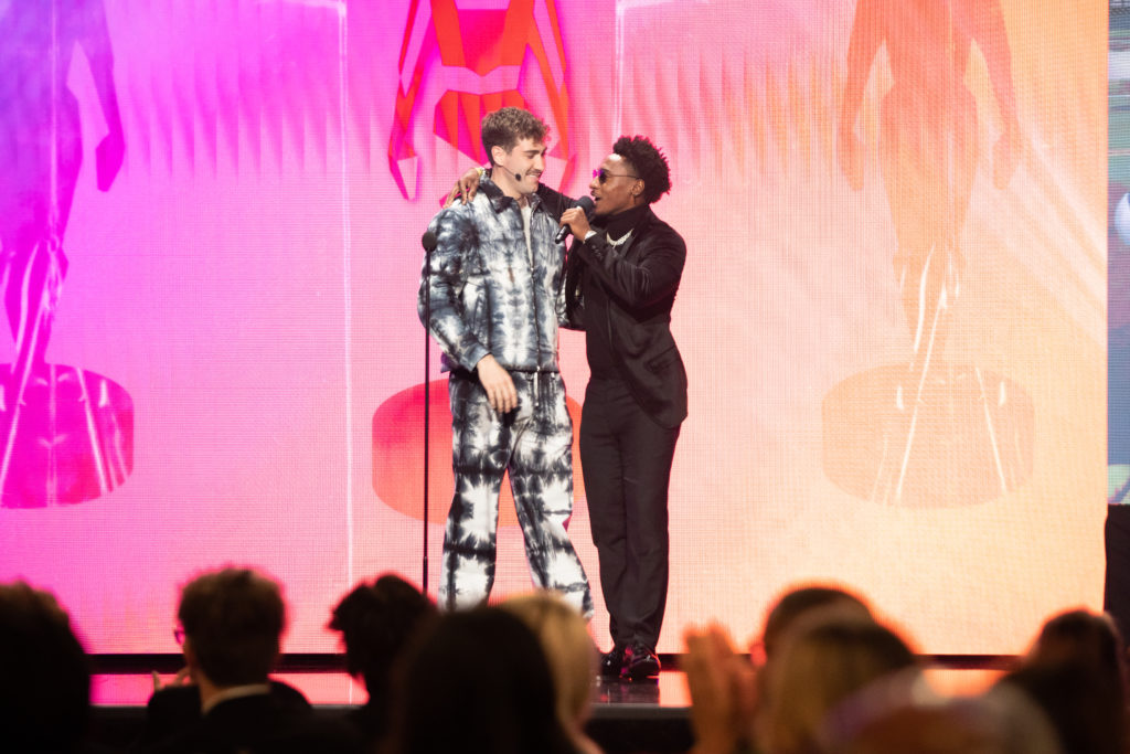 Creator FaZe Deestroying hugging Airrack and speaking into a microphone on stage of the Streamy Awards with a pink and orange background
