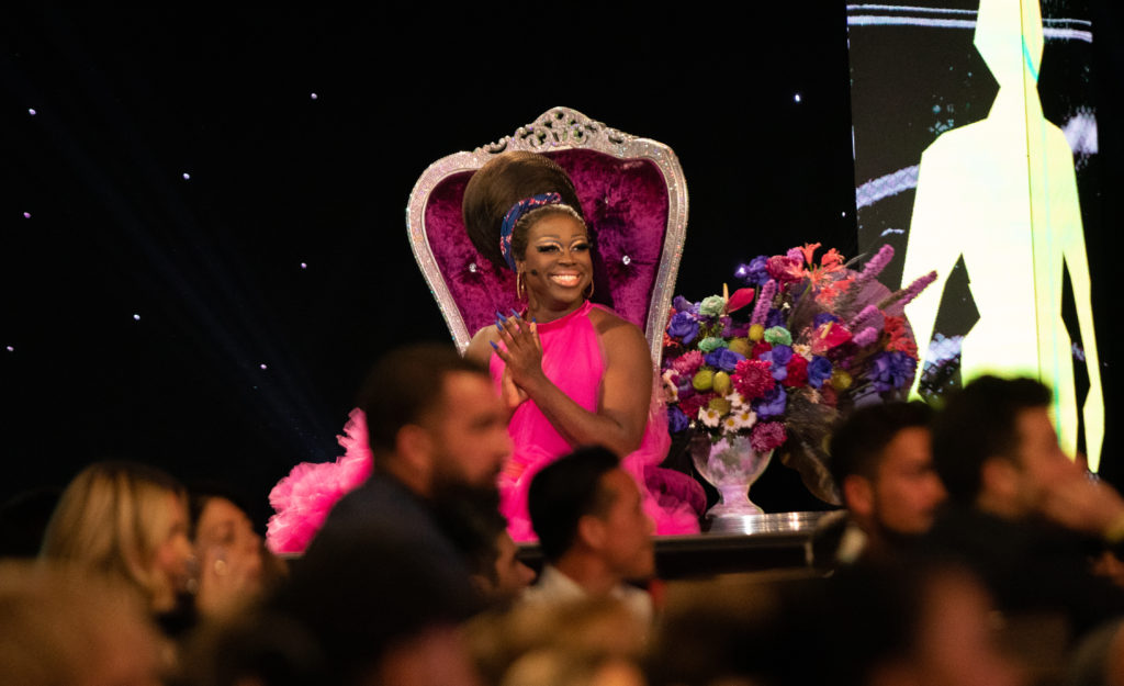 Bob the Drag Queen sitting on stage in a pink chair wearing a pink dress smiling and clapping looking out into the audience
