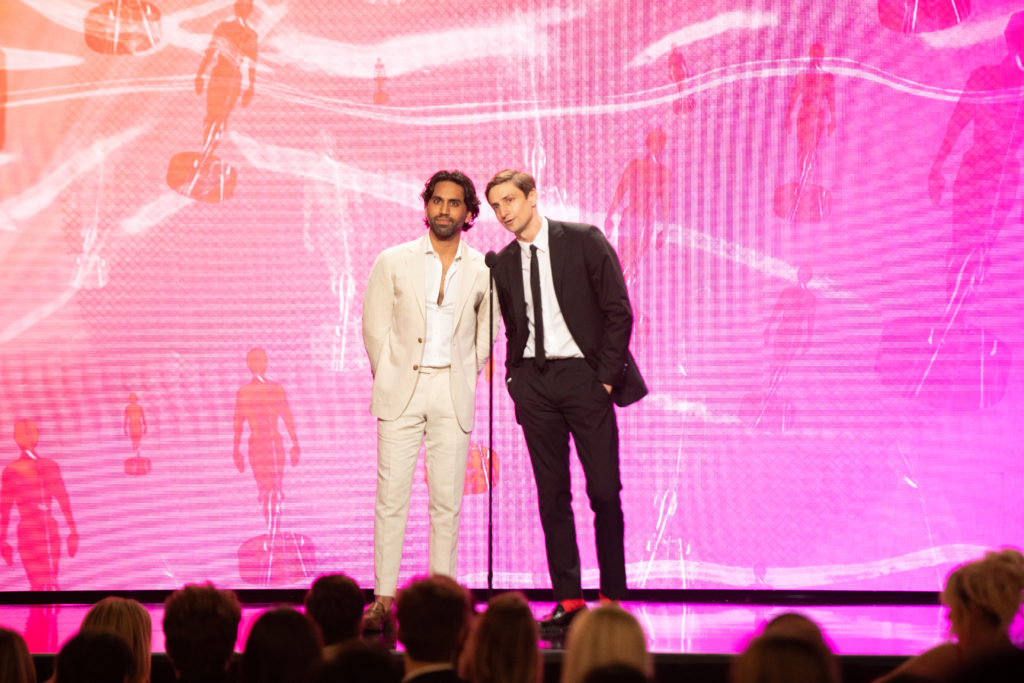 Creators Colin and Samir standing on stage at the Streamy Awards in front of a pink and orange background talking into a microphone