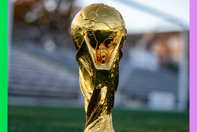 2022 World Cup trophy sitting on a soccer field in front of a goal and stadium seating with a rainbow gradient border.