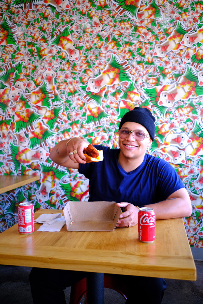 Creator SpicyyCam sitting at a table in front of a colorful printed wall smiling holding a sandwich.