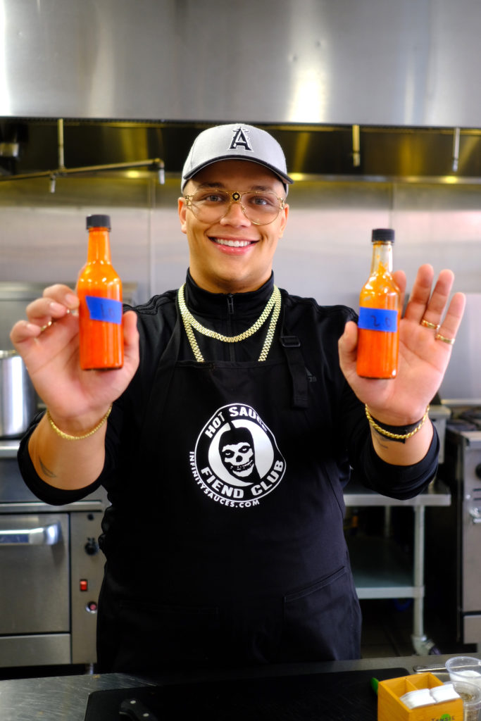 Creator SpicyyCam standing in a kitchen smiling at the camera holding two bottles of hot sauce in each hand.