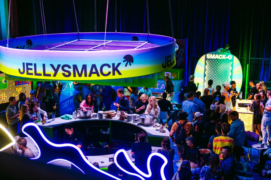 Round Jellysmack sign hangs over a large round desk in the middle of a crowded party surrounded by neon lights