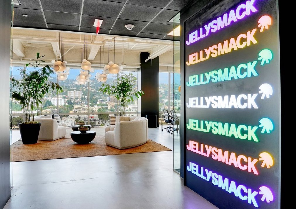 Repeating Jellysmack logo neon sign in a rainbow of colors next to a view of an office setting with couches, plants, lighting and a window looking out into the Hollywood Hills